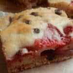 Close-up of a strawberry chocolate chip bar on parchment, cut to show the jammy strawberry layer beneath a golden top