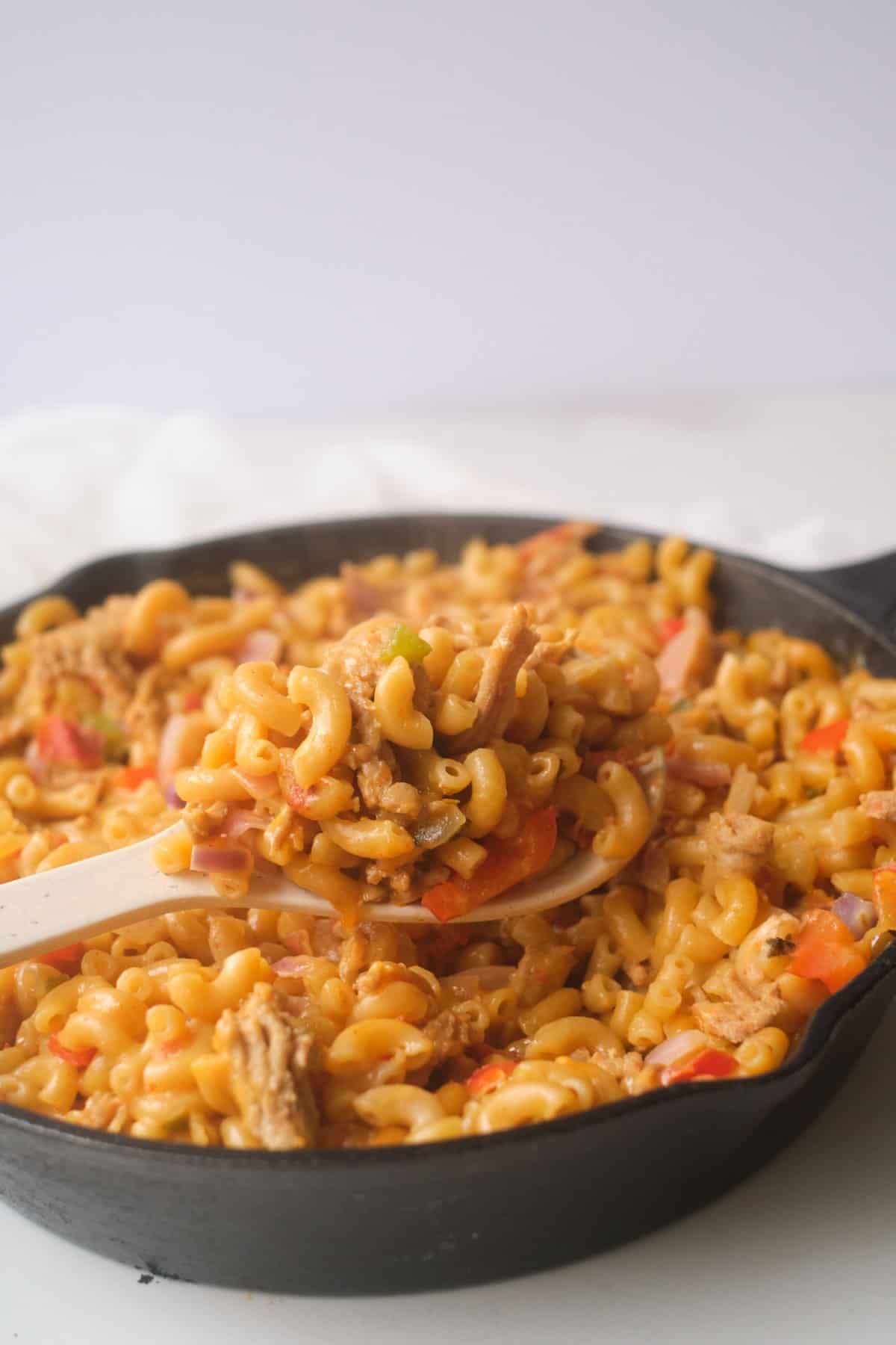 Wooden spoon lifting chicken taco mac and cheese from a cast iron skillet, elbow macaroni in an orange sauce with shredded chicken, diced red pepper and red onion