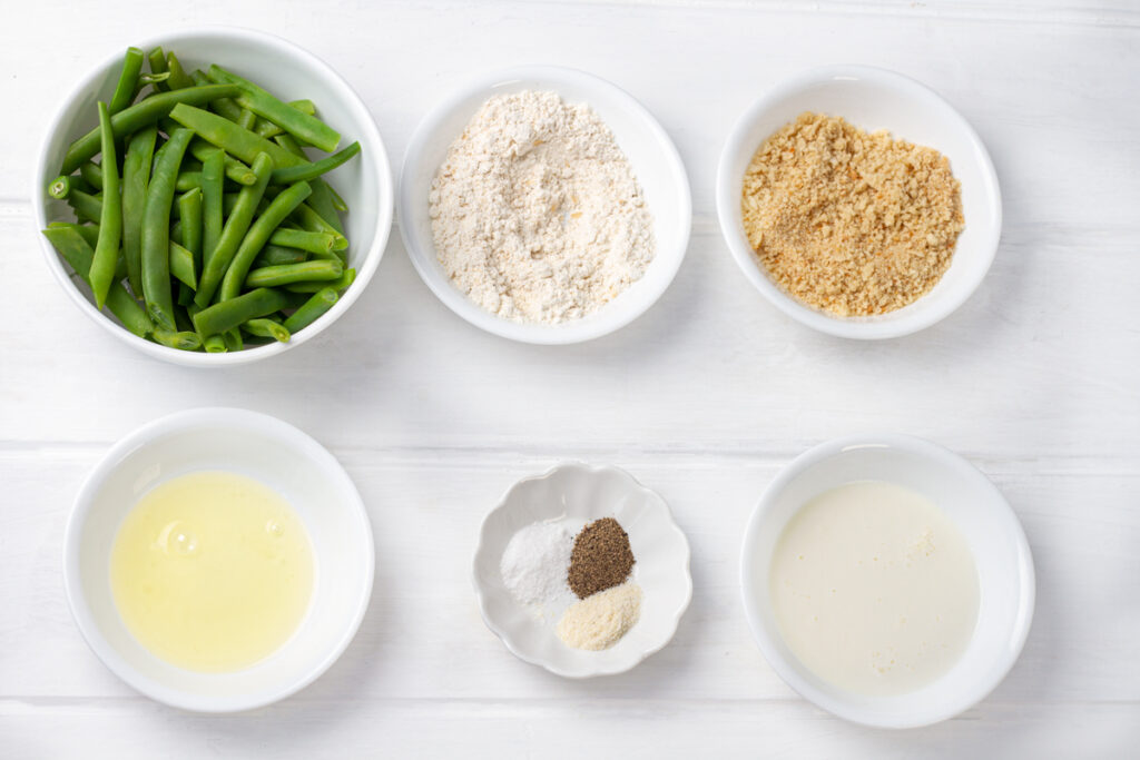 A dish of green beans, a dish of flour, a dish of Panko bread crumbs, a dish of egg whites, a dish of spices, and a dish of milk. 