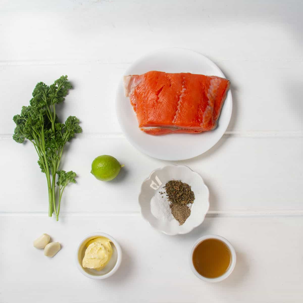Raw salmon fillet, a bunch of parsley, a lime, seasonings in a dish, softened butter, two garlic cloves, and cornstarch dissolved in broth. 