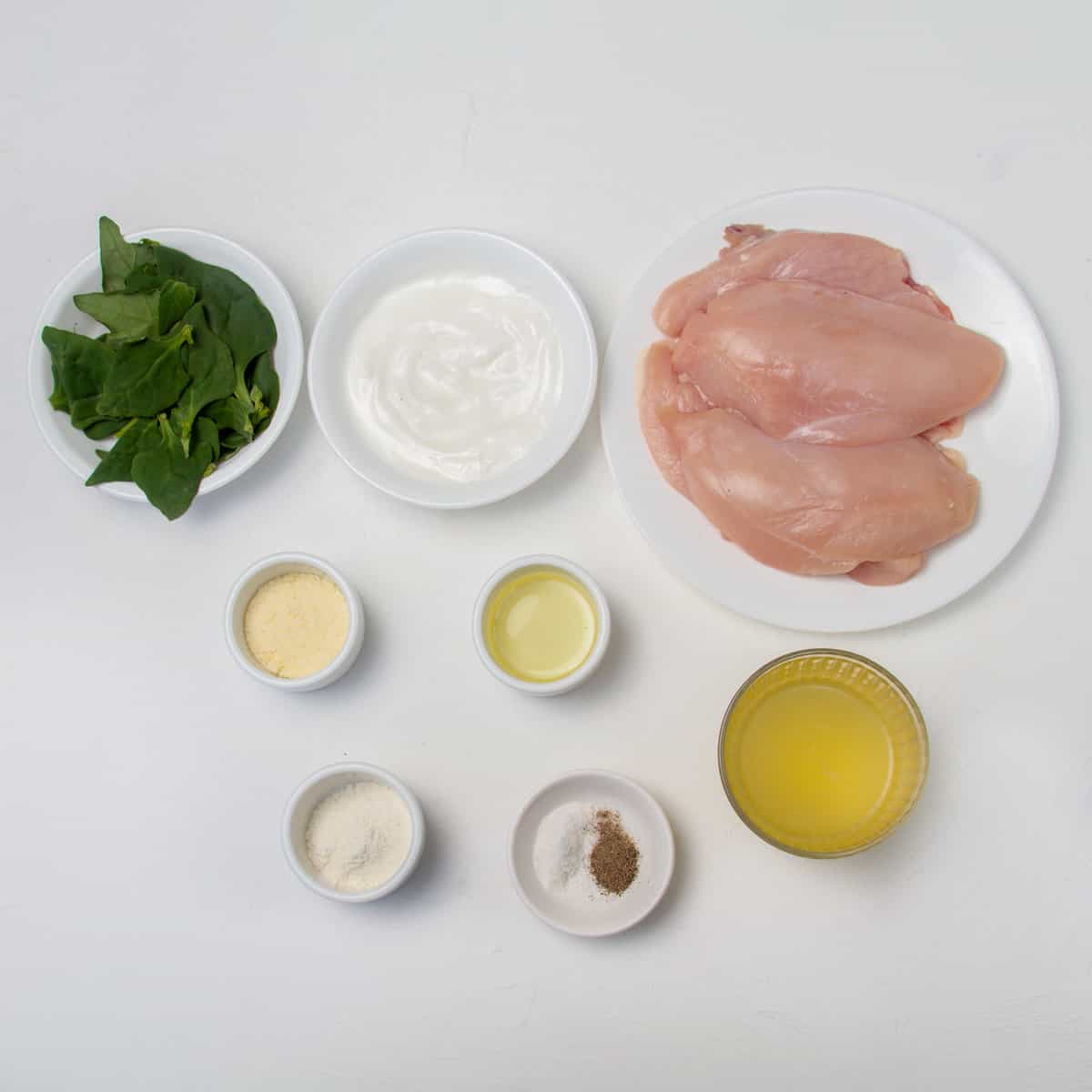 Raw chicken breast, yogurt, spinach, oil, parmesan cheese, cornstarch, salt, and pepper in separate dishes. 