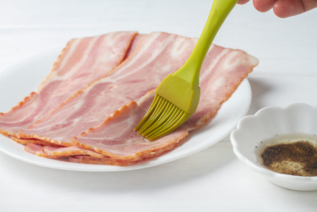 A hand using a silicone brush to coat turkey bacon in oil and spice mixture. 