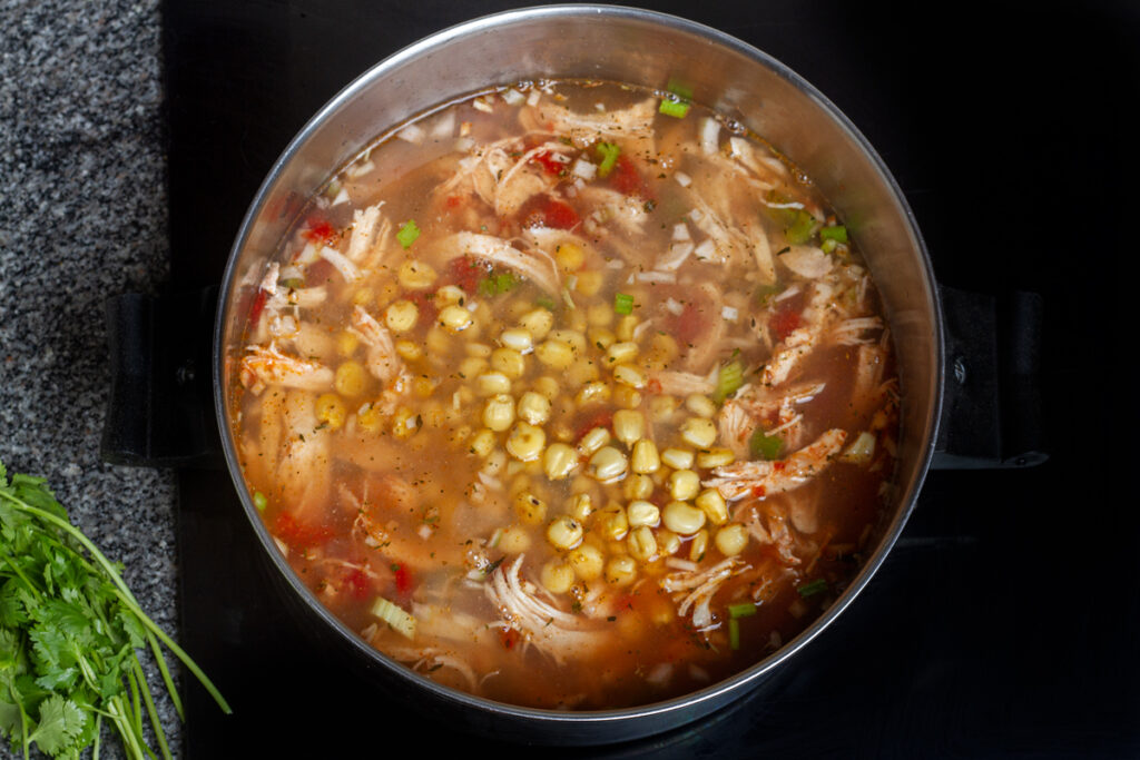 Shredded chicken, celery, corn, and tomatoes cooking in a clear broth. 