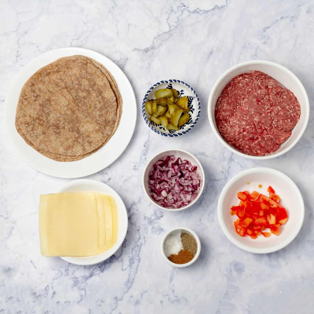 Smashed taco ingredients of tortillas, raw ground beef, peppers, onions, pickles, tomatoes, cheese, and seasonings in separate dishes.
