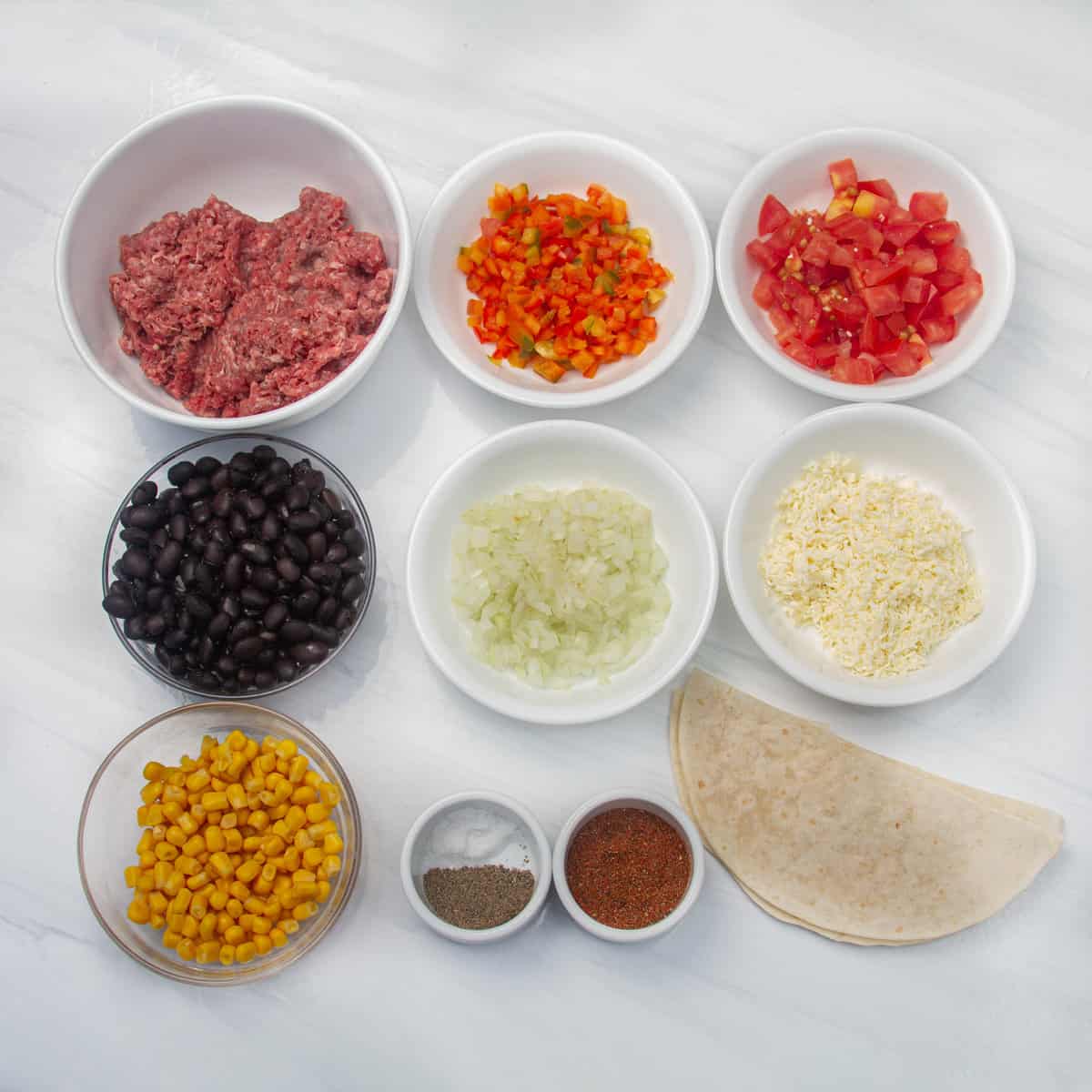 Ingredients of ground beef, taco seasoning, black beans, sweet corn, diced tomatoes, diced onions, chopped bell pepper, grated cheese, tortillas, and Greek yogurt in separate dishes. 