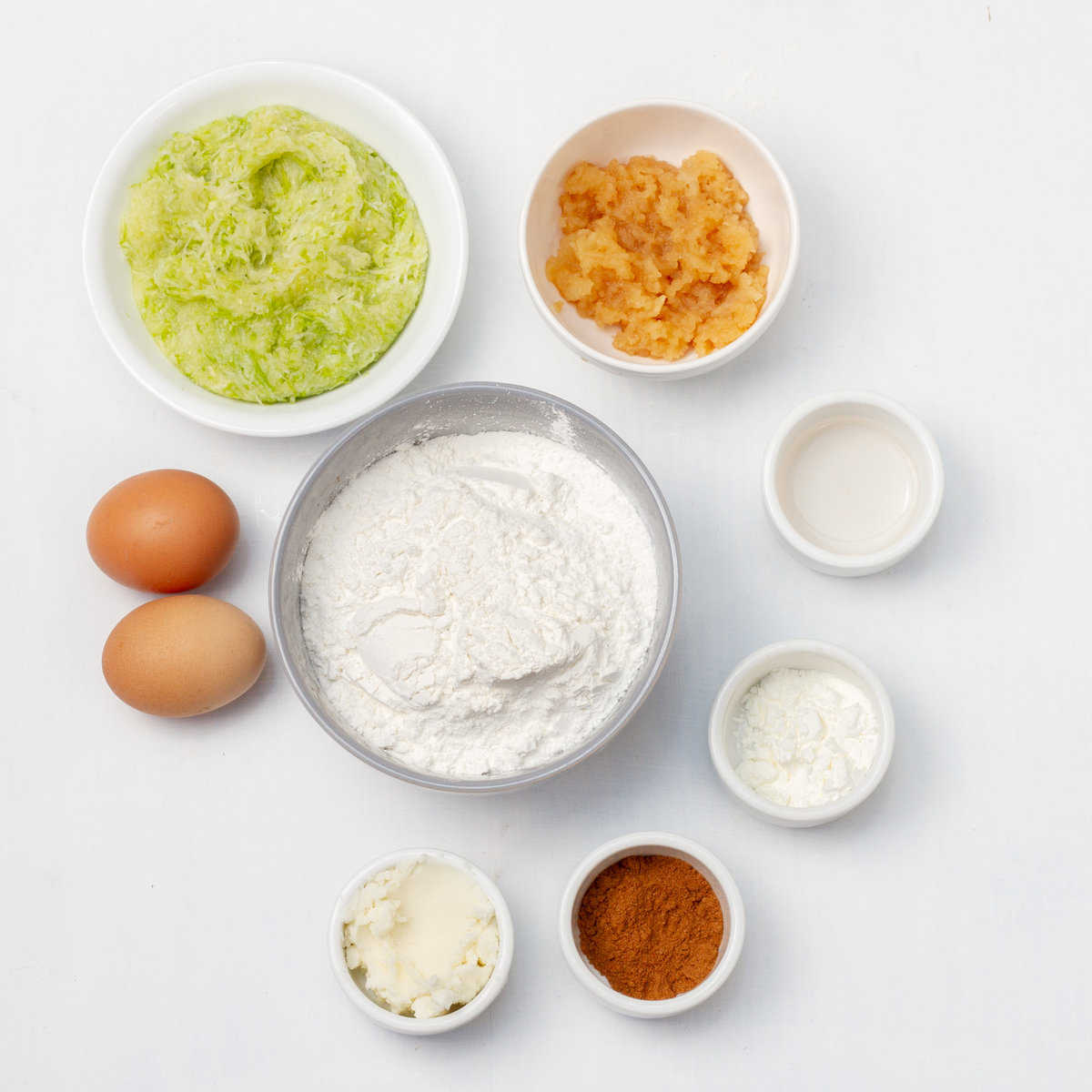 Ingredients of gluten-free all-purpose flour, baking powder, ground cinnamon, grated apples, stevia, coconut oil, vanilla, grated zucchini, and salt in separate dishes. 