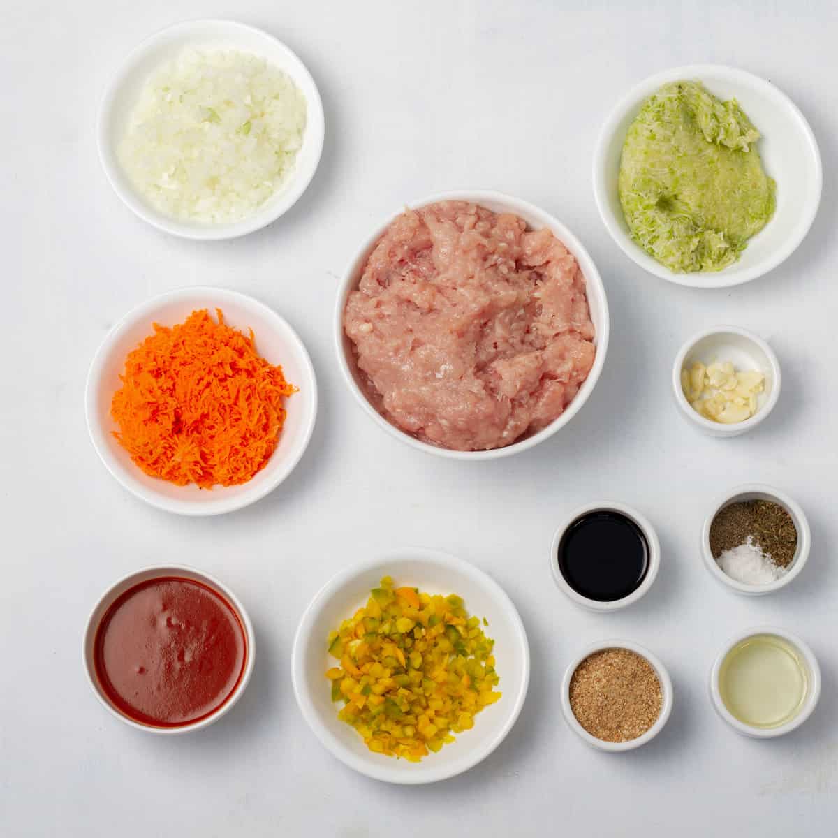 Ingredients of ground turkey, breadcrumbs, onion, garlic, carrot, zucchini, egg, sauce, and spices in separate dishes.