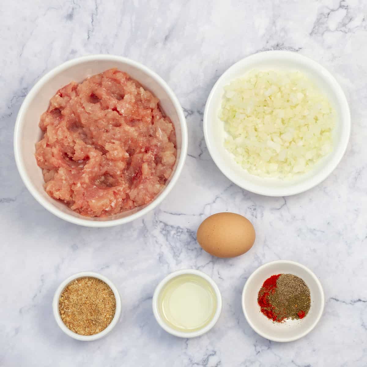 Ingredients of raw ground chicken breast, diced onion, spices, and an egg in separate dishes.