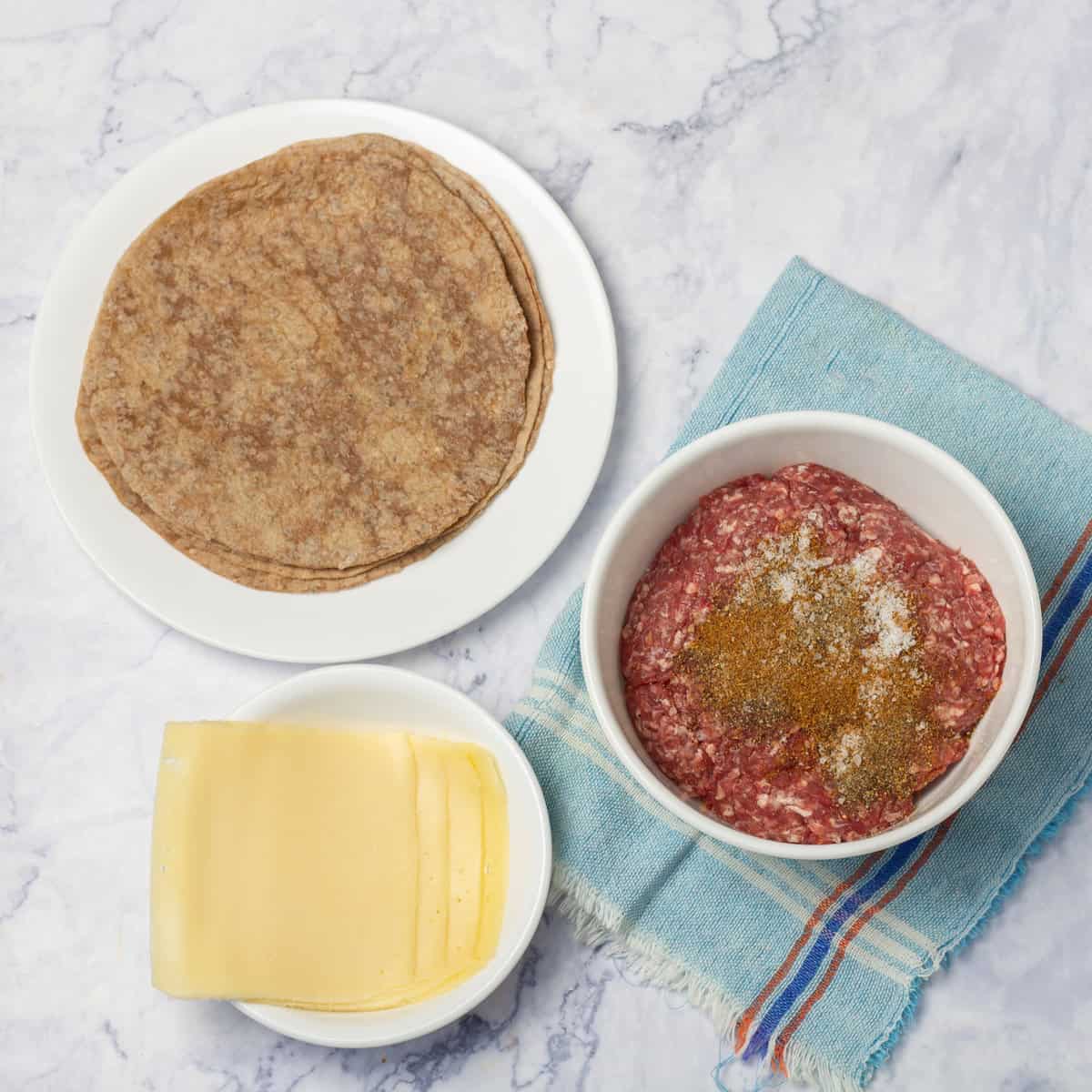 Ground beef marinating in seasonings with tortillas and cheese in separate dishes.