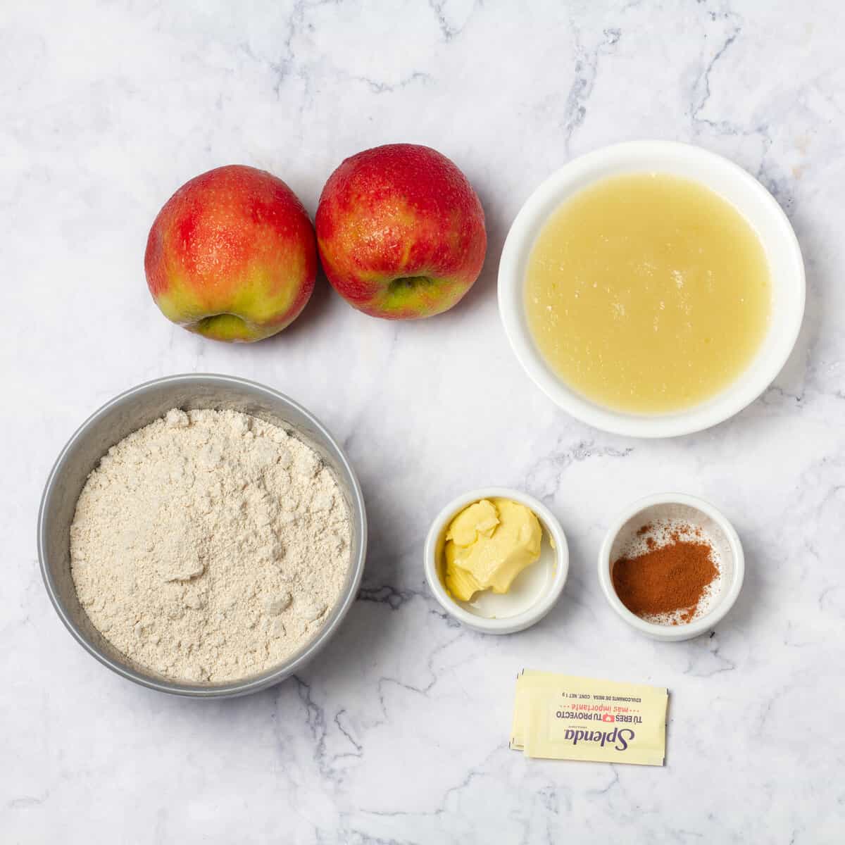 Ingredients of apples, oat flour, butter, sweetener, and spices in separate dishes. 