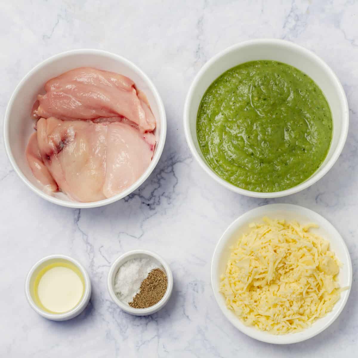 Ingredients of raw chicken breast, salsa verde sauce, mozzarella cheese, and seasonings in separate dishes. 