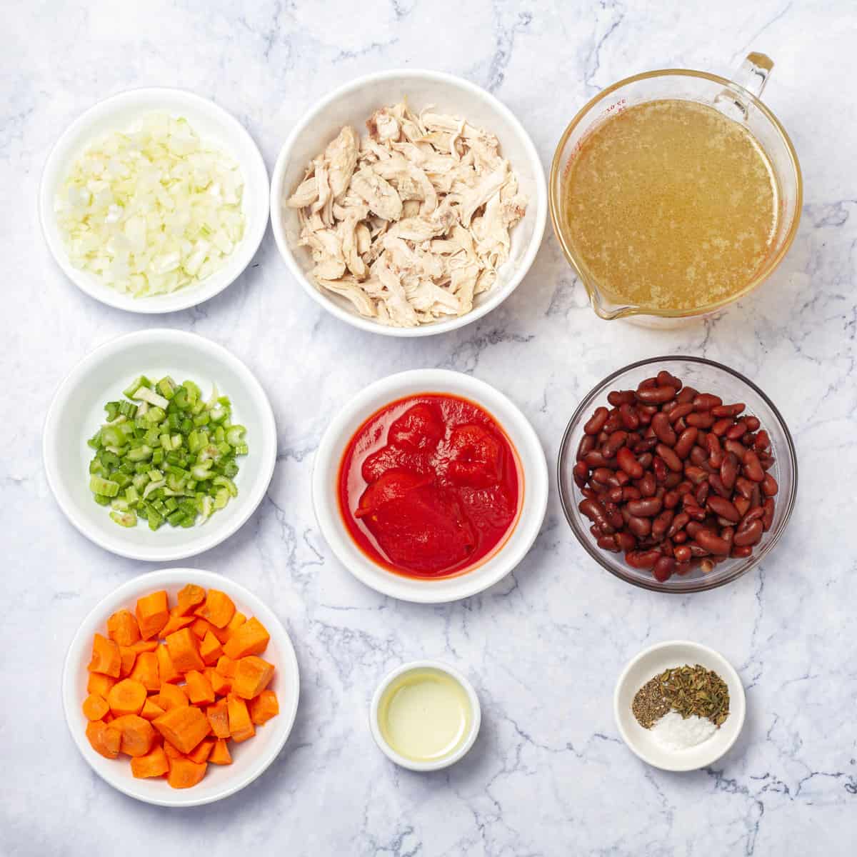 Ingredients of shredded chicken, kidney beans, onions, tomatoes, carrots, chicken broth, and spices in separate dishes. 