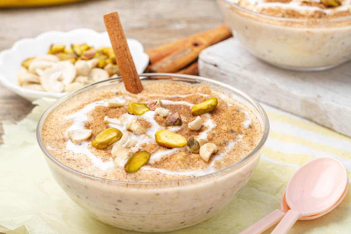 A bowl of overnight cinnamon roll oats on a table with a cinnamon stick in it and spoons.