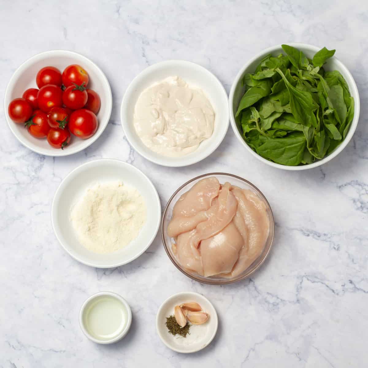 Tuscan chicken ingredients of chicken breast, spinach, cherry tomatoes, fat-free Greek yogurt, parmesan cheese, Herbs de Provence, garlic, and salt in separate dishes. 