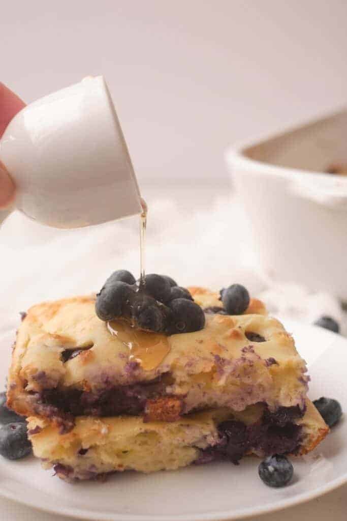 A square of baked blueberry lemon pancake on a plate with syrup being poured on it.