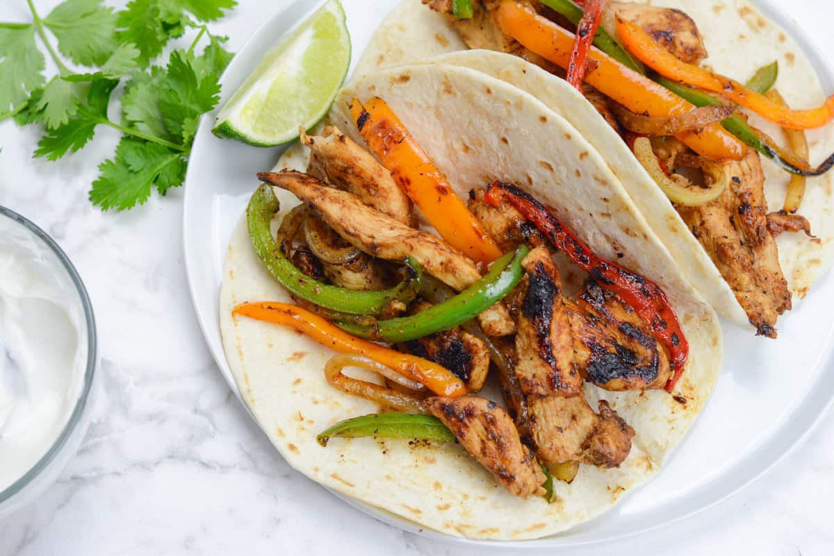 Two tortillas filled with cooked fajita chicken, peppers and onions sit on a white plate. A lime wedge sits to the side of the fajitas. Fresh cilantro leaves and a small bowl of sour cream sit to the left of the plate. 