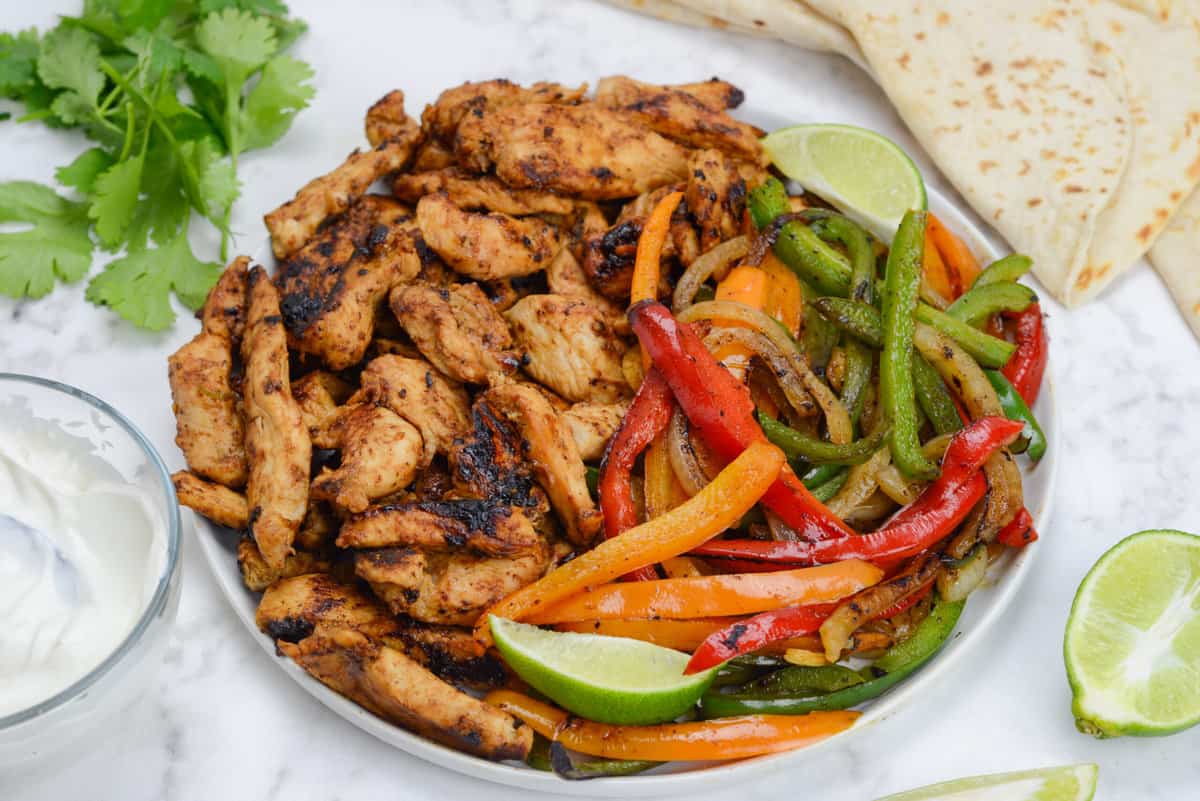Large plate of charred fajita chicken and red, orange and green bell peppers. Slices of lime garnish the sit and sit to the right. Tortillas, cilantro and a small bowl of sour cream surround the plate.