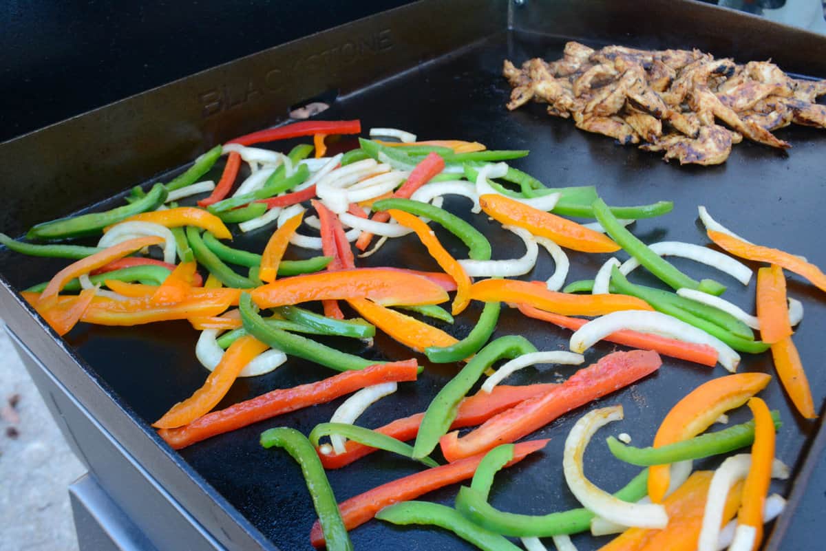 Orange, red and green pepper slices and white onion slices sit on the left side of the Griddle. In the background the chicken can be seen scooped together on the right side of the griddle. 
