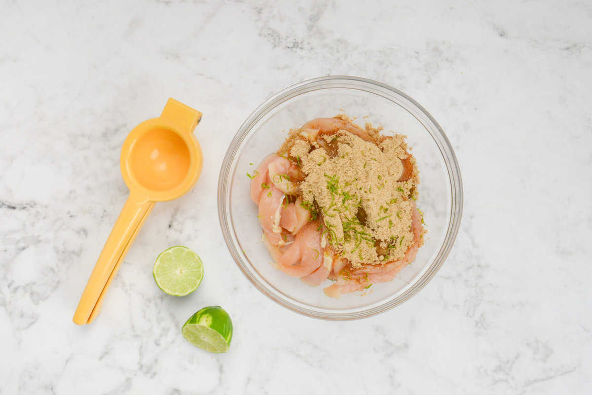 A bowl filled with sliced chicken is topped with fajita seasoning and fresh lime zest. To the left is a hand held juicer and a lime cut in half.