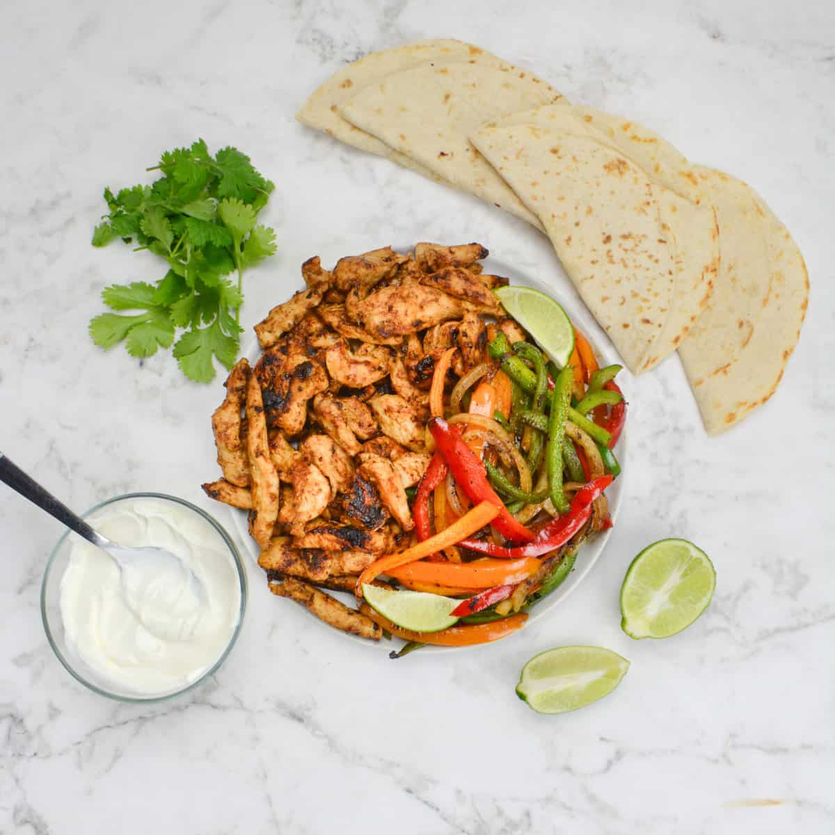 A plate of cooked fajita chicken and an assortment of peppers and onions sit on a white plate. Tortillas, cilantro, a halved lime and a small bowl of sour cream surround.