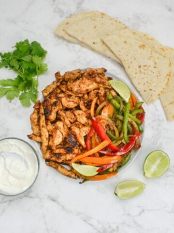 A plate of cooked fajita chicken and an assortment of peppers and onions sit on a white plate. Tortillas, cilantro, a halved lime and a small bowl of sour cream surround.