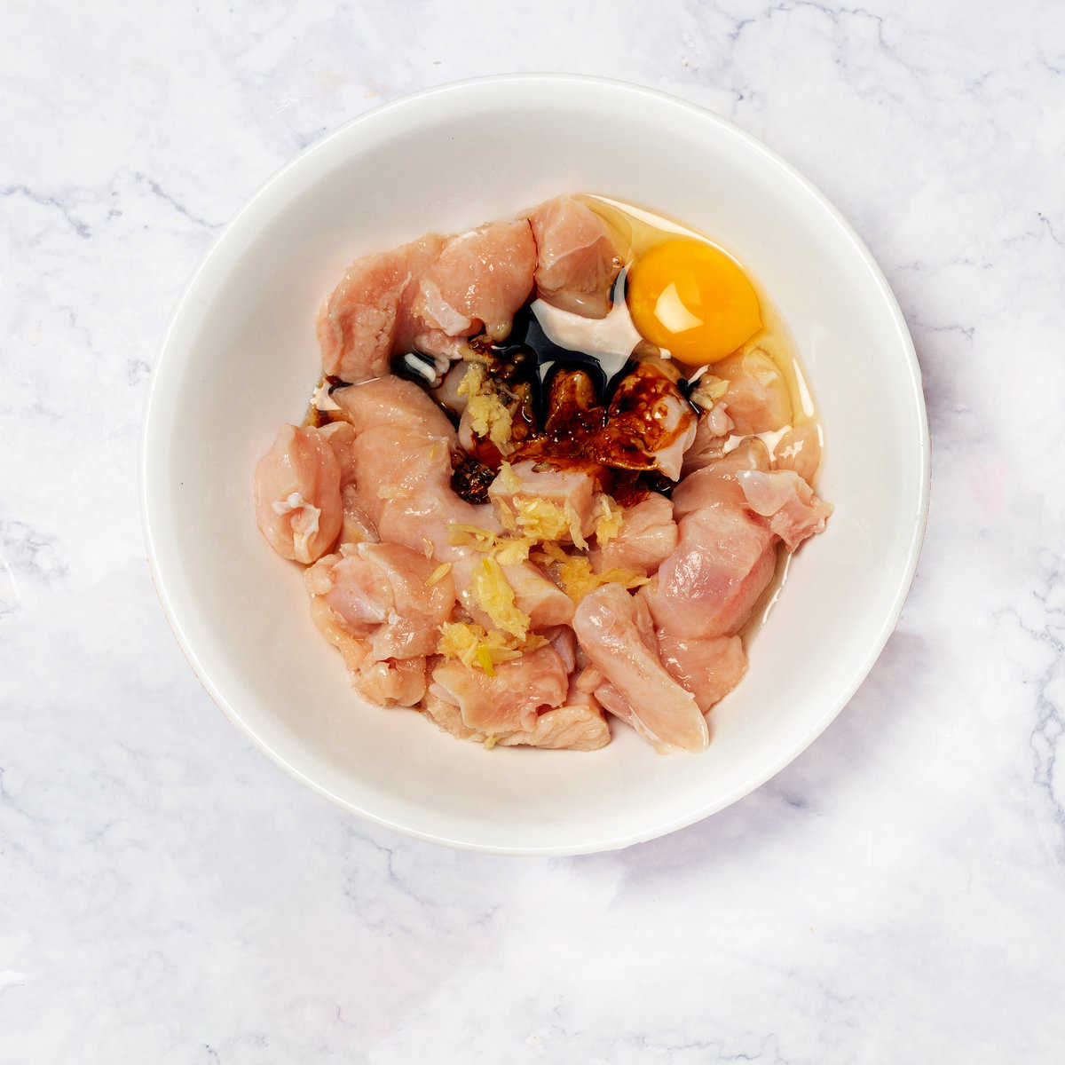 A bowl of chicken cubes with crushed garlic, soy sauce, a beaten egg, and pepper