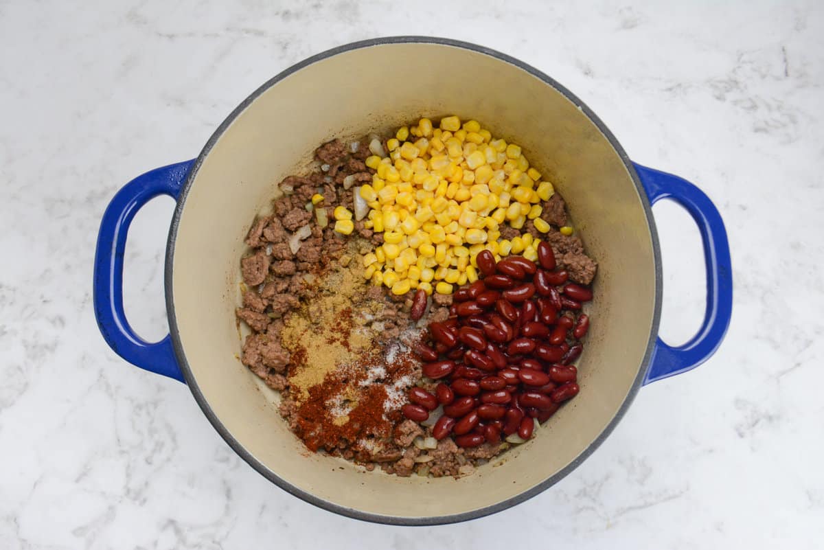 A blue Dutch oven is shown with corn, beans and seasoning now added to the pot.