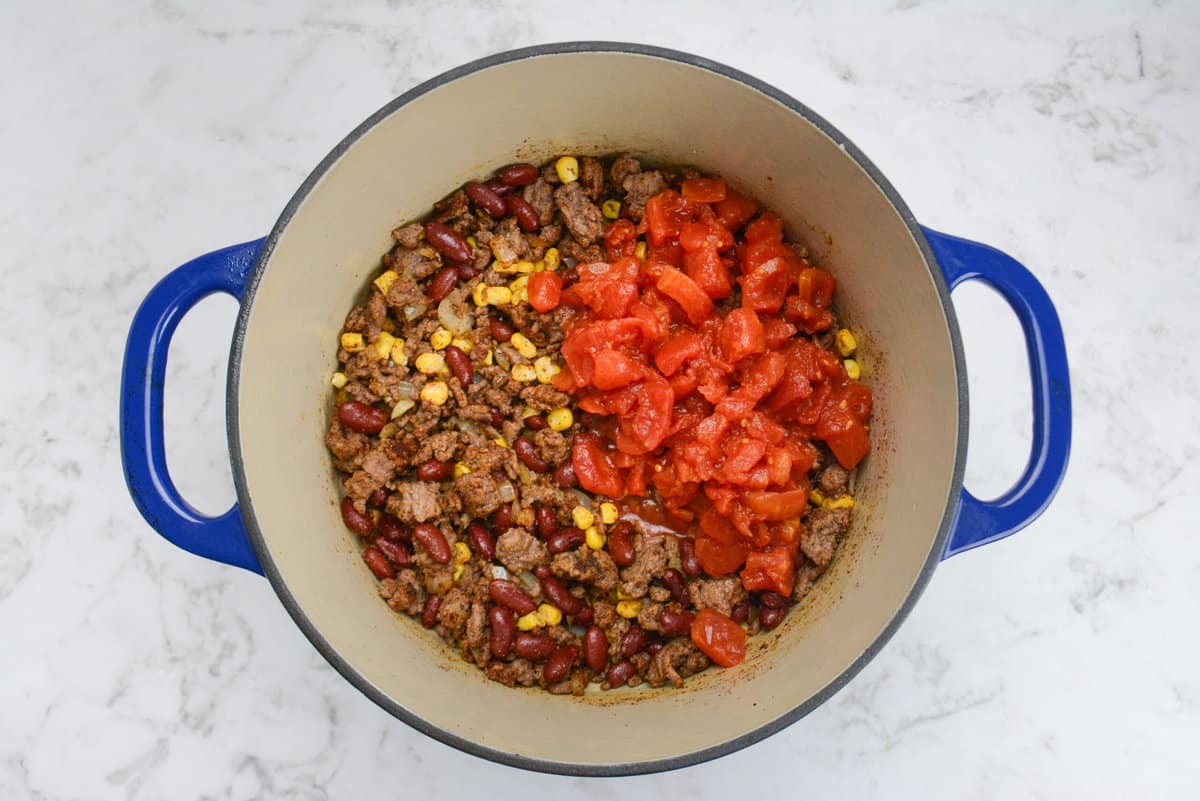 A blue Dutch oven is shown with the mixed beef mixture and vibrant tomatoes on top.
