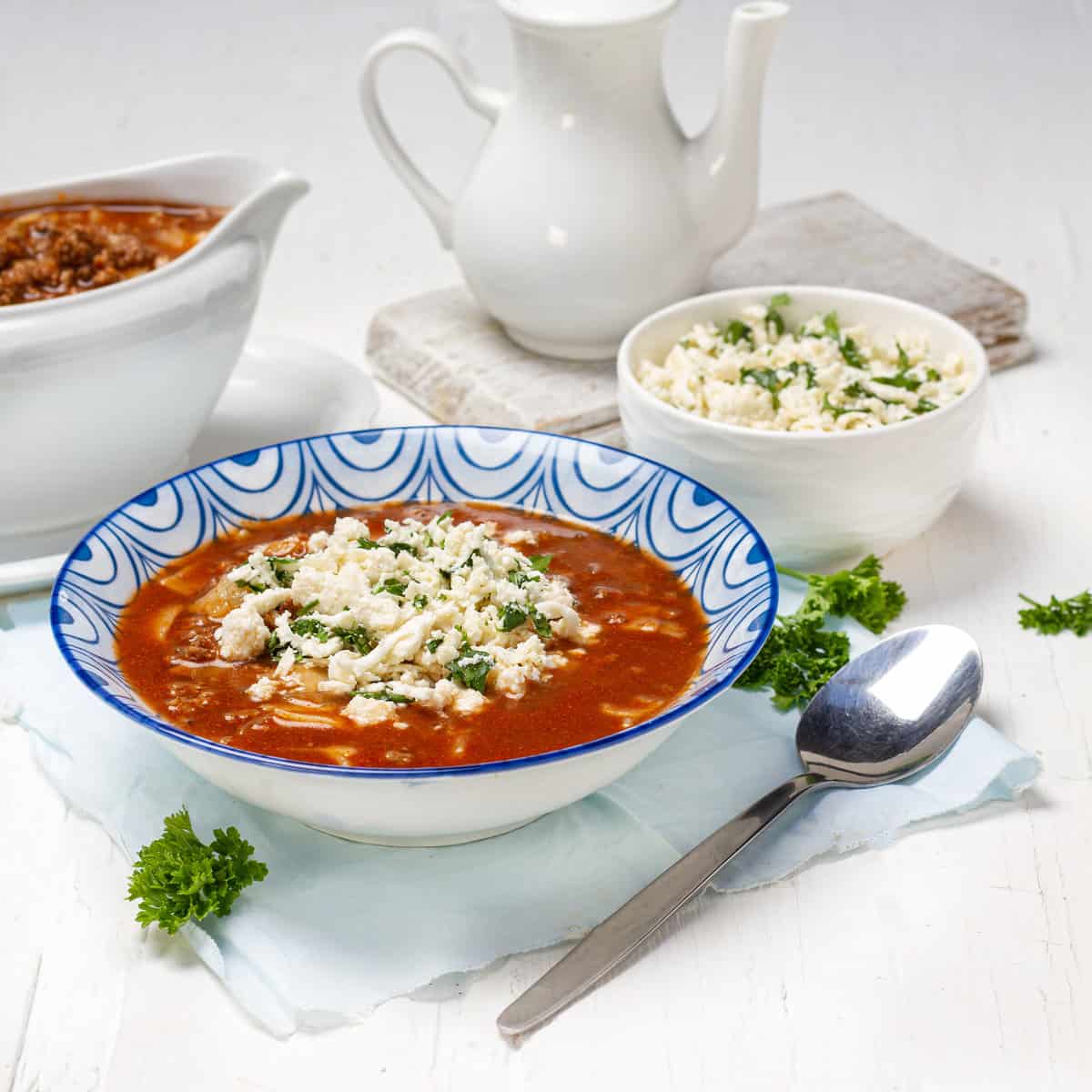 blue and white bowl of red lasagna soup with broken noodles, topped with crumbled cheese and parsley, a white pot and a bowl of cheese behind