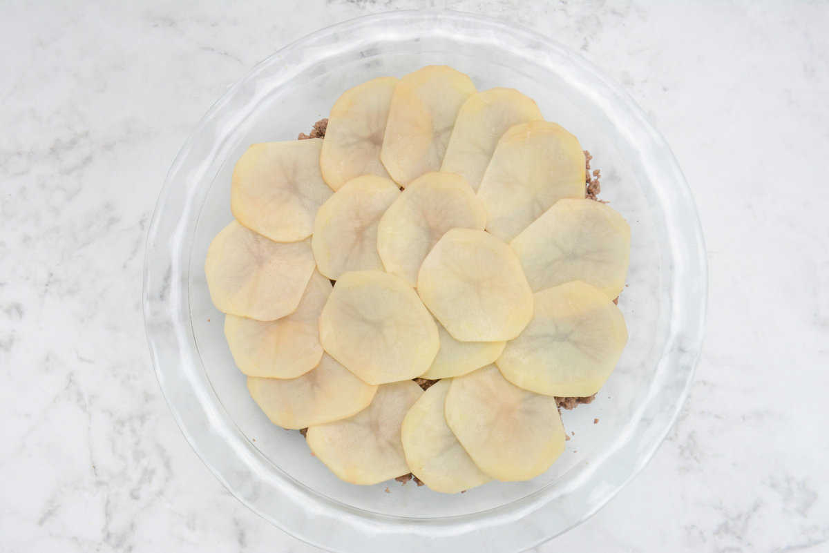 A clear pie plate is shown with potato slices overlapping the ground beef 
