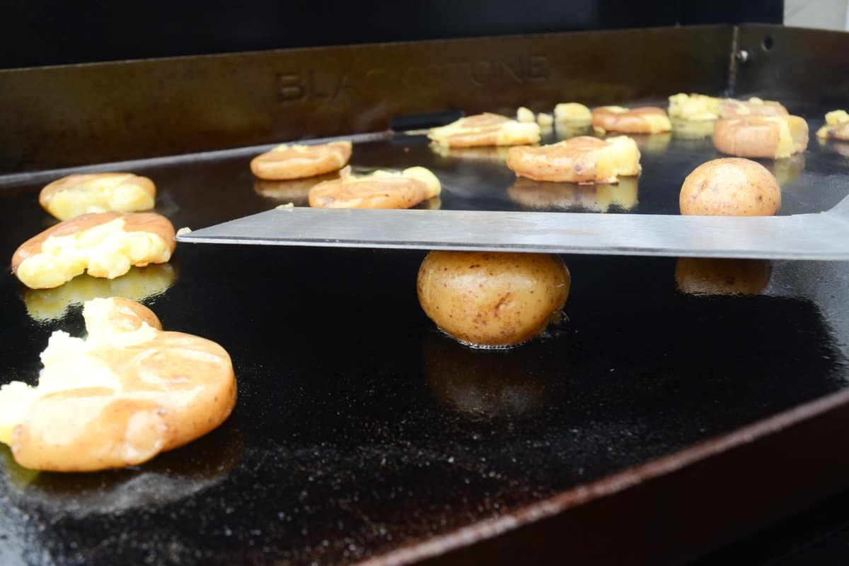 freshly smashed potatoes sitting on a blackstone griddle. a spatula is smashing one potato in the foreground.