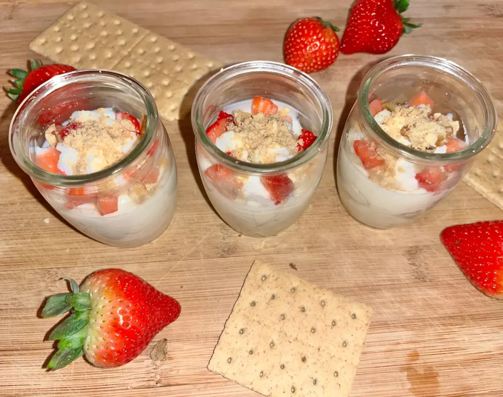Three mugs of strawberry cheesecake cups on a table with graham crackers and strawberries on a table.