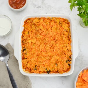 A white square baking dish filled filled with Doritos casserole is surrounded by small bowls filled with salsa, Greek yogurt and Doritos. A serving spoon and brown napkin sit to the side.