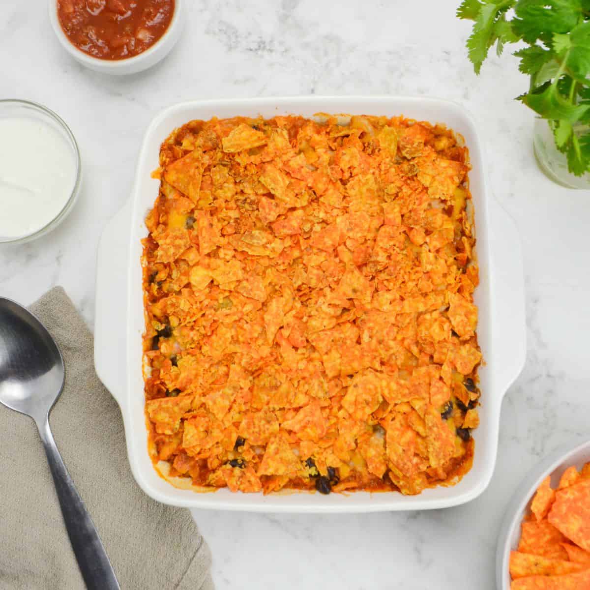 A white square baking dish filled with Doritos casserole sits on a white table. Surrounding it are small bowls of Greek yogurt, salsa, cilantro, and Doritos. A serving spoon on a brown napkin sits to the side. 