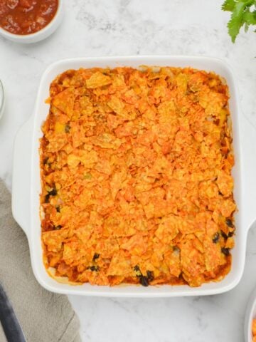 A square white baking dish filled with Doritos Casserole is shown. A serving spoon on a napkin sits to the side. Small bowls with Greek Yogurt, salsa and Doritos surround the dish.