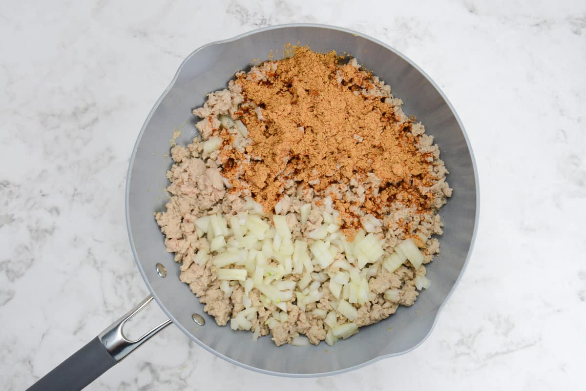 A skillet with cooked light brown ground chicken, taco seasoning and diced onions.