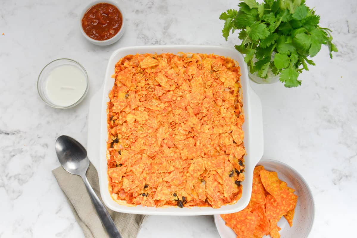 A white square baking dish is shown with the now cooked casserole. To the side are a serving spoon, bunch of cilantro and bowls filled with Doritos, Greek Yogurt and salsa. 