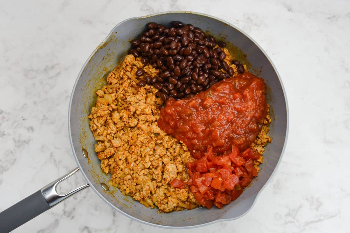 A skillet with ground chicken, black beans, salsa, and diced tomatoes that are not mixed together.
