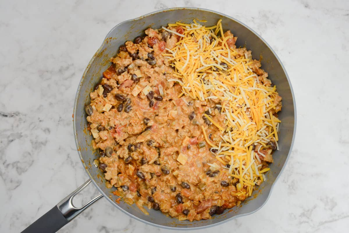 A skillet is shown with the casserole ingredients mixed well with a portion of orange and white cheese on top.