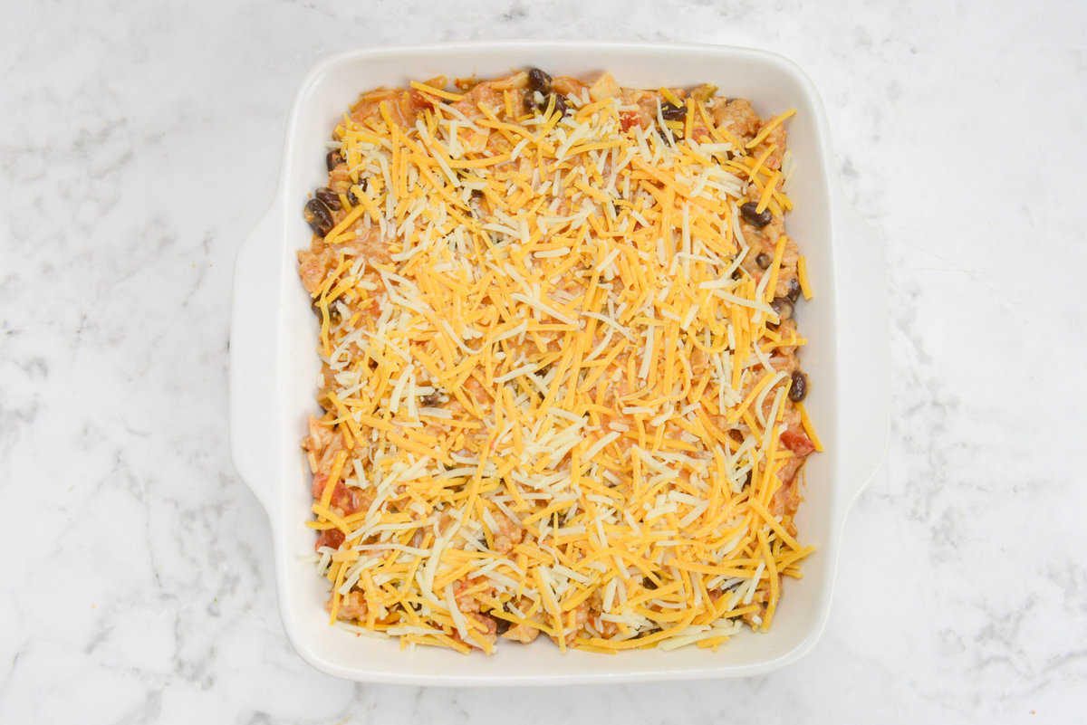 A white square baking dish filled with the casserole mixture and topped entirely with orange and white shredded cheese.