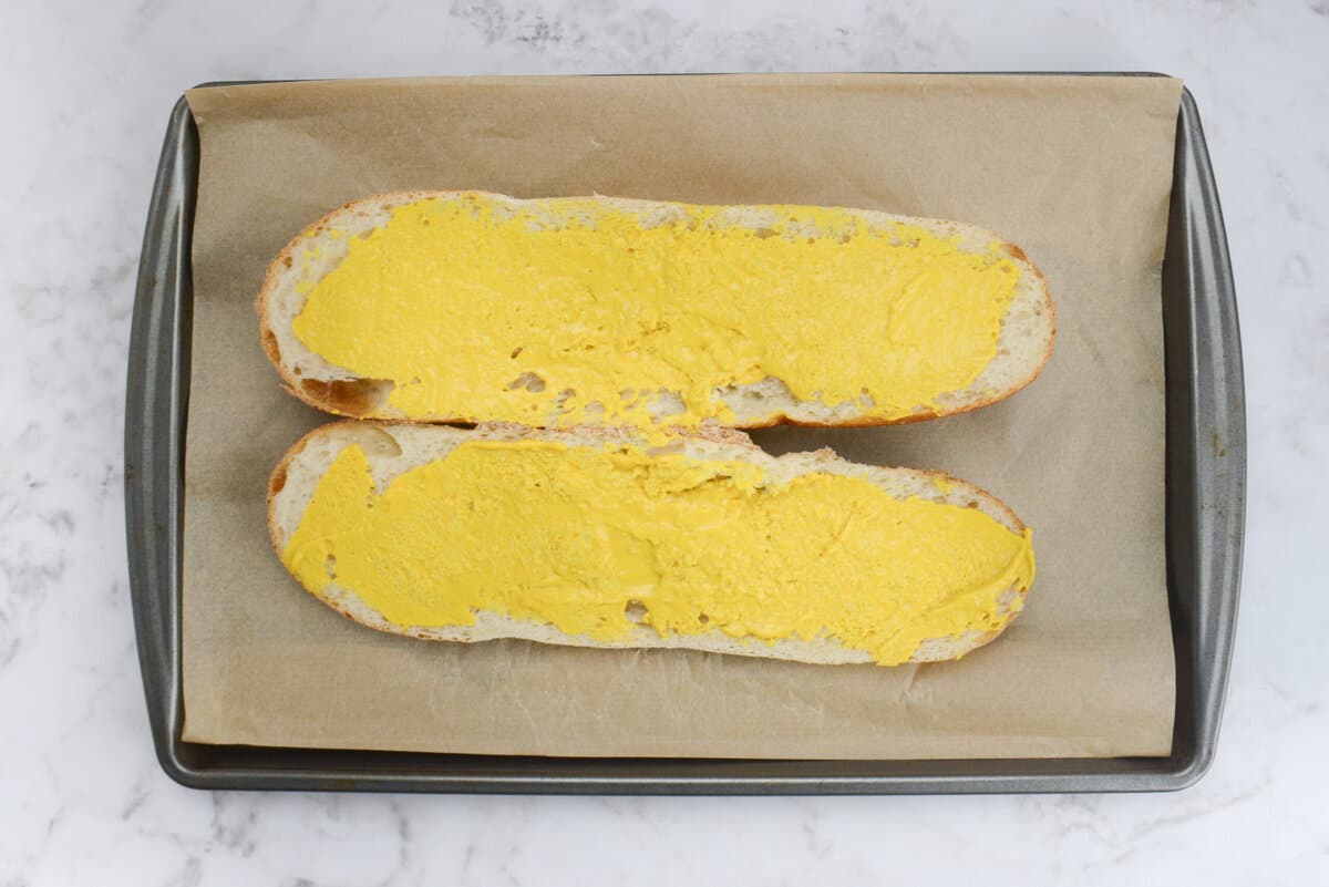 Sliced French bread on parchment paper with mustard generously spread on both sides. 