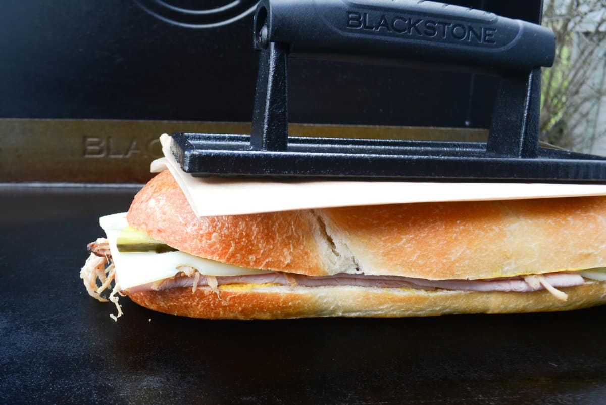 Cuban sandwich cooking on Blackstone griddle, being pressed down by griddle press. 