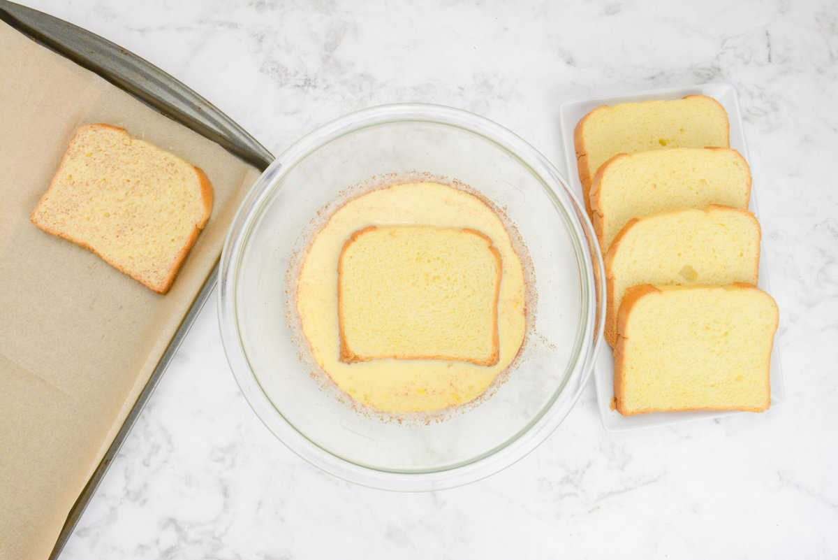 A slice of bread is placed into a large mixing bowl filled with the custard mixture. A slice of dipped bread sits to the left while four slices of undipped bread are placed to the right.