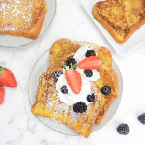 A plate filled with three slices French toast topped with whipped cream, berries, and powdered sugar. Two plates and berries sit to the side.