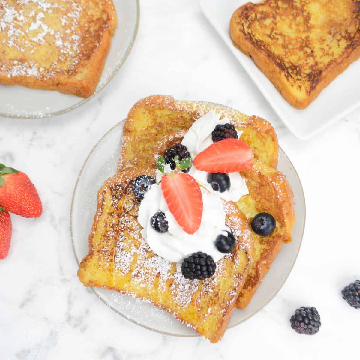 Three slices of Blackstone French toast sit on a plate topped with whipped cream, powdered sugar, berries and honey. Two plates of untopped French toast are placed to the side.