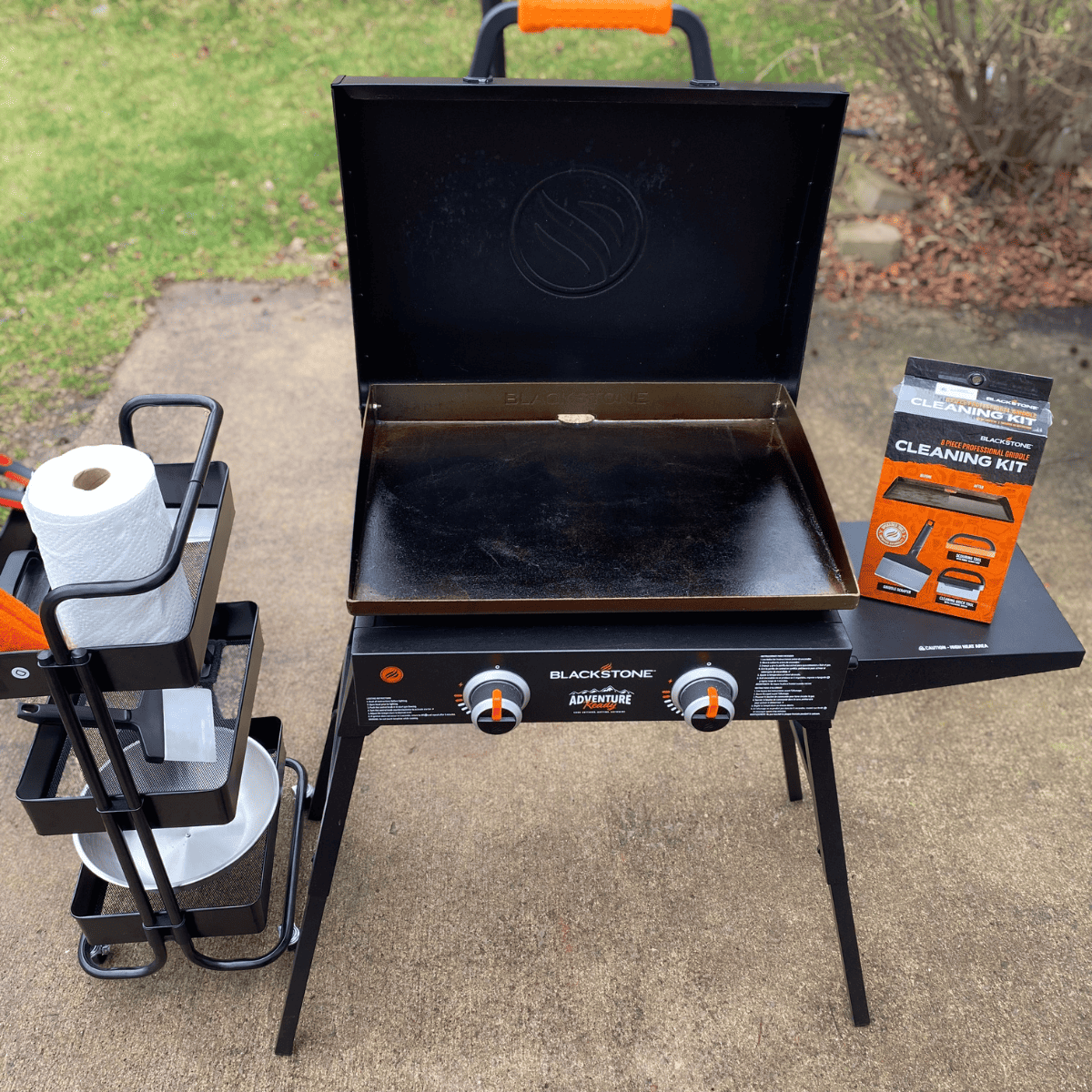 Blackstone griddle on patio with lid open. Cleaning kit sits on the side table while an accessory cart is to the right of the griddle