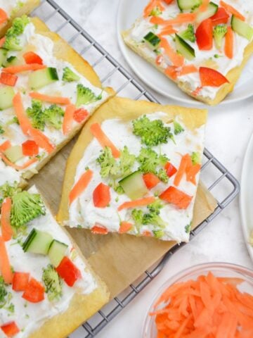 Four slices of cold veggie pizza are on a cooling rack lined with parchment. A small plate with one slice sits off to the side. A small bowl of shredded carrots is below.