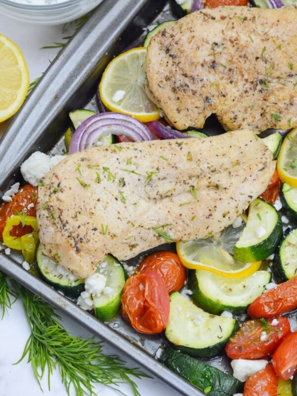 Close up of cooked Greek sheet pan chicken. Two pieces of chicken are on top of roasted vegetables in a sheet pan. A cut lemon and sprigs of dill are to the side.