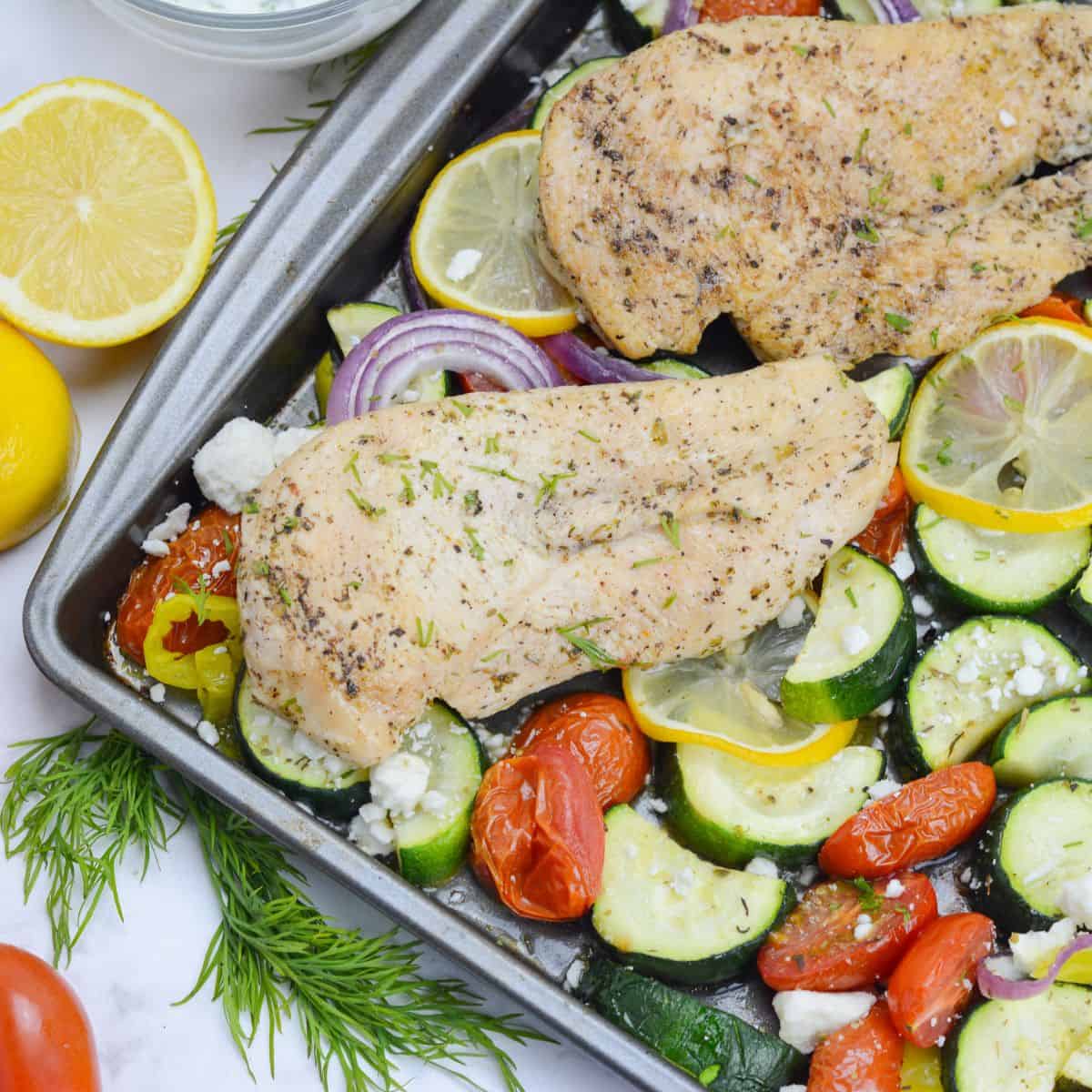 Close up of cooked Greek Sheet Pan chicken. Two pieces of chicken are placed on top of roasted vegetables a sheet pan. A cut lemon and sprigs of dill are to the side. 