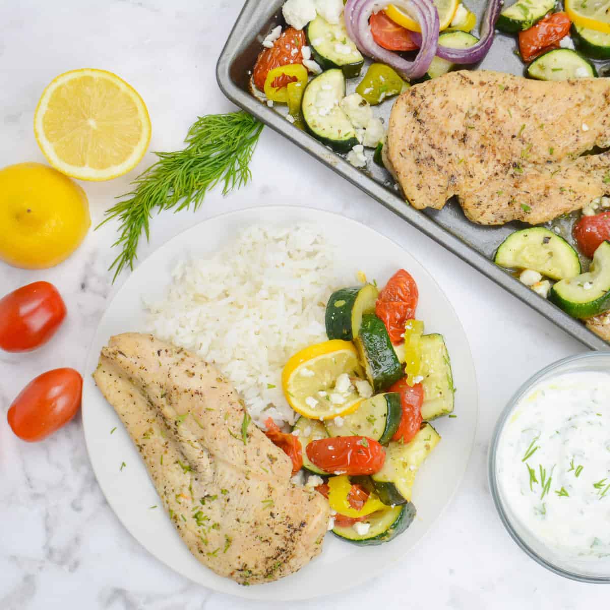 A white plate with a serving of chicken, roasted vegetables and white rice.To the side sits the sheet pan with additional servings, a bowl of tzatziki, a cut lemon, dill and tomatoes. 